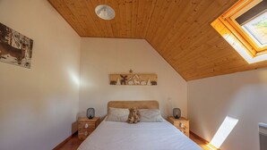 Une chambre avec un plafond en pente, une lucarne, un lit avec une literie blanche, un ours en peluche posé sur les oreillers et un élément décoratif fixé au mur au-dessus du lit.