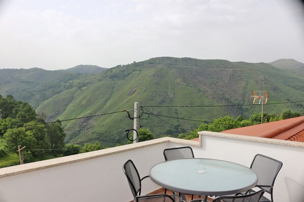 A balcony with a round glass table and four chairs, overlooking a mountainous landscape.
