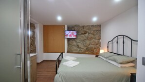 A bedroom with a bed, a television mounted on the wall, a wooden wardrobe, and a stone wall. - House in Gerês for 2 people with fireplace (Gerês)