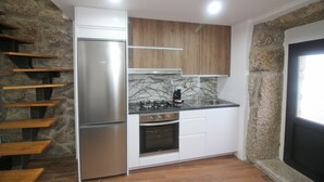 A compact kitchen with a built-in refrigerator, oven, and gas stove. It features a stone accent wall and wooden cabinets. - House in Gerês for 2 people with fireplace (Gerês)