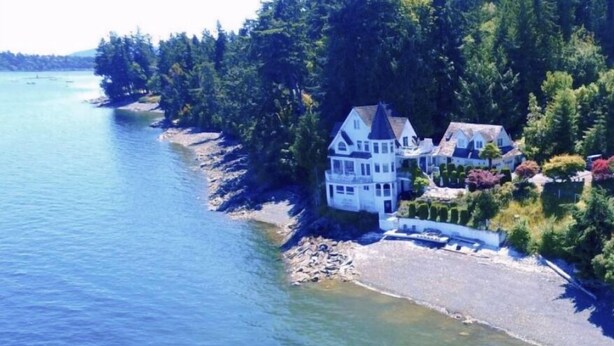 A coastal property with a large house and multiple smaller buildings, surrounded by dense forest and a body of water.
