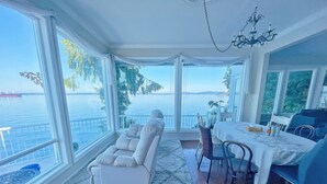 A room with a large window overlooking a body of water, a white sofa, a dining table with chairs, and a chandelier.