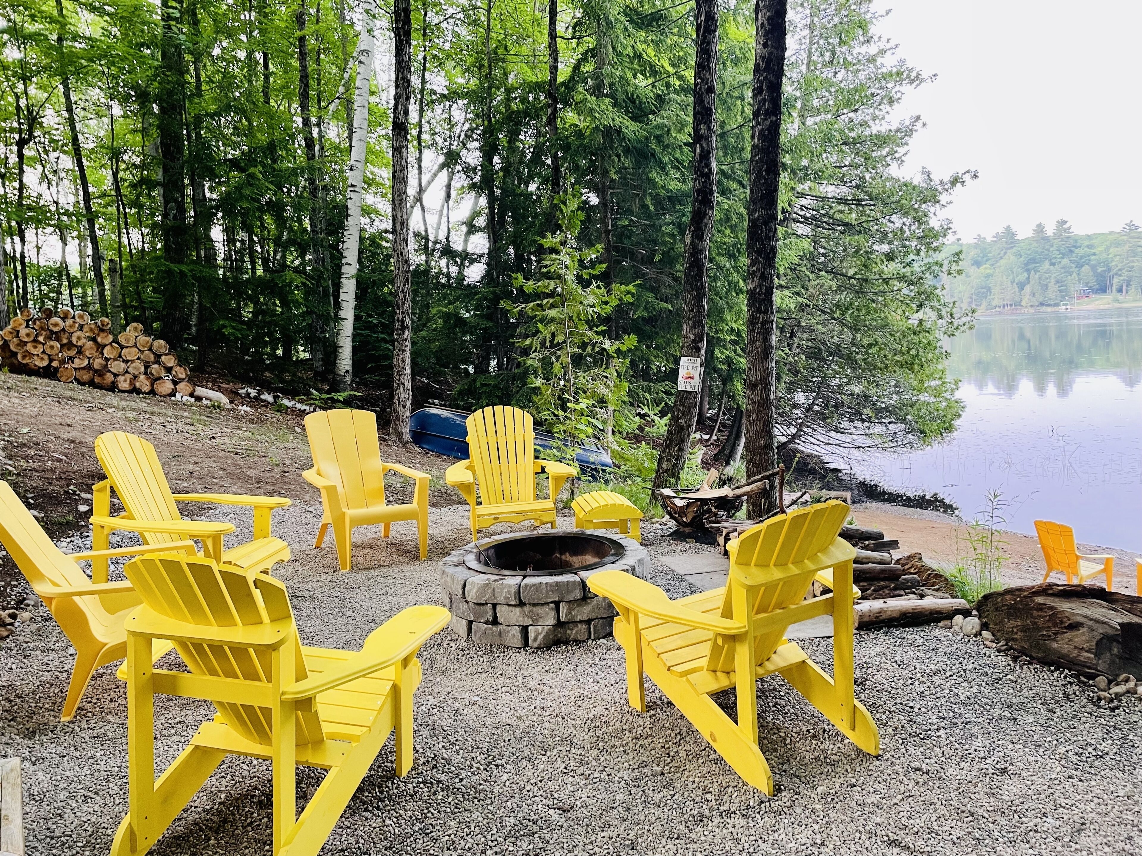 A lakeside seating area with yellow Adirondack chairs arranged around a stone fire pit.