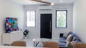 A modern living room with a flat-screen TV, a round coffee table, a sofa, and a dining area with chairs. There are two windows with blinds, a door, and a ceiling light fixture. - CENTRALLY-LOCATED CONDO (San Juan)