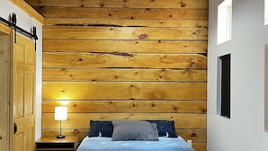 A bedroom with a wooden slat wall, a bed with a star-patterned bedspread, a bedside table with a lamp, and a window with blinds.