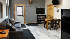 A living room with a gray couch, a wooden dining table with chairs, a mounted deer head, a bookshelf, and a TV on the wall.