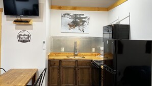 A compact kitchen with a wooden table, chairs, a black refrigerator, a sink, and a mounted TV.
