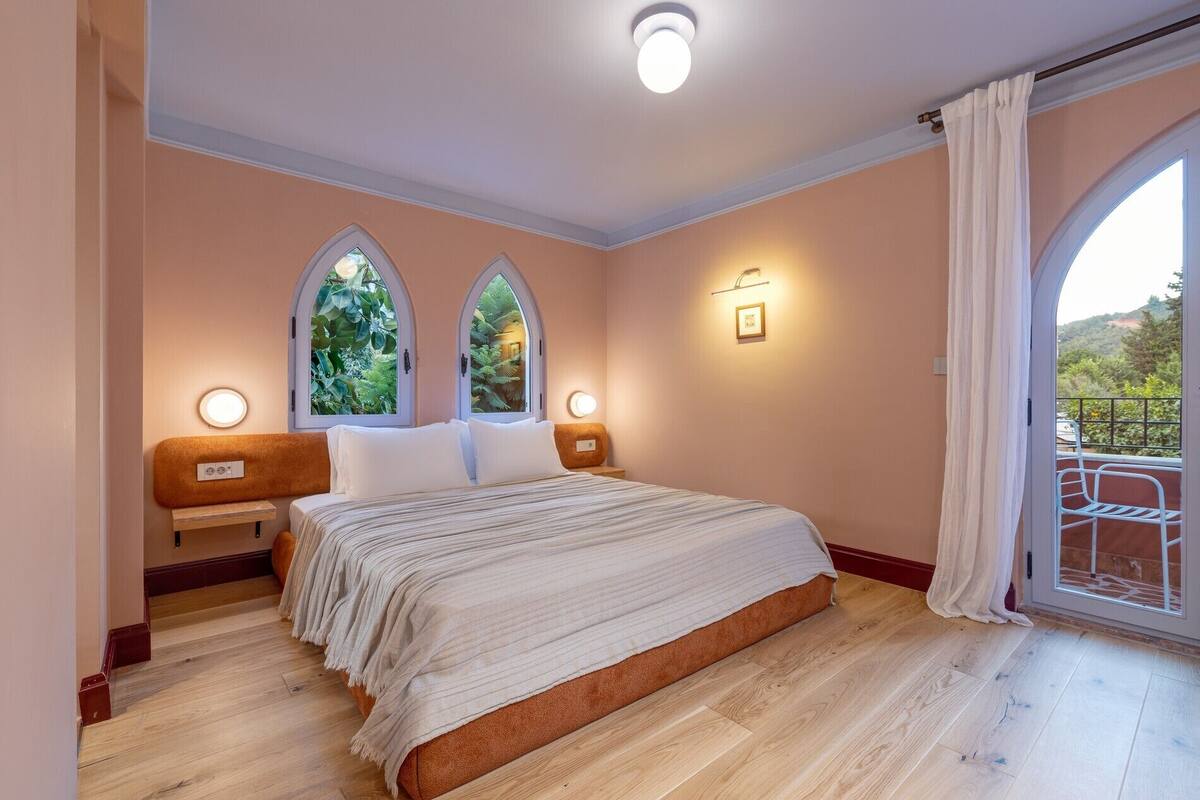 Une chambre avec un grand lit, deux tables de chevet, une commode, une lampe fixée au mur et une fenêtre donnant sur un paysage verdoyant.