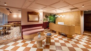 A hotel lobby with a checkered floor, a reception desk, a seating area with a pink sofa, and a large mirror on the wall. - Yonca Anjelika Otel (Fethiye)