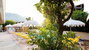 A poolside area with sun umbrellas, lounge chairs, and a tree in the foreground. - Yonca Anjelika Otel (Fethiye)