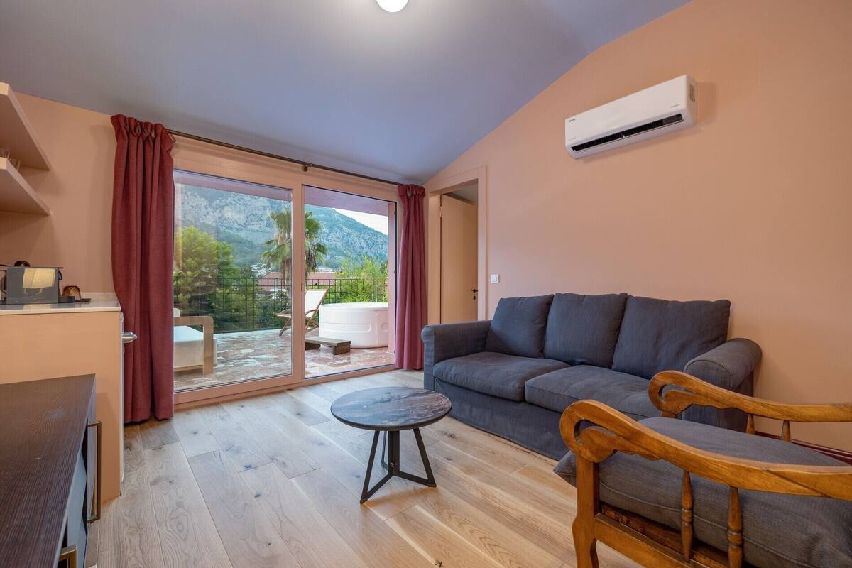 Un salon moderne avec un canapé gris, des chaises en bois, une petite table ronde et une porte-fenêtre coulissante donnant sur un balcon avec vue sur la montagne.