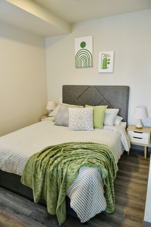 A neatly made bed with striped bedding, a green throw blanket, and decorative pillows. There are two bedside tables with lamps and a small bedside table with a lamp. The room has a modern design with a grey headboard and a wooden floor. - Cozy loft perfect for home office and relax. (Nunciatura, Rohrmoser)