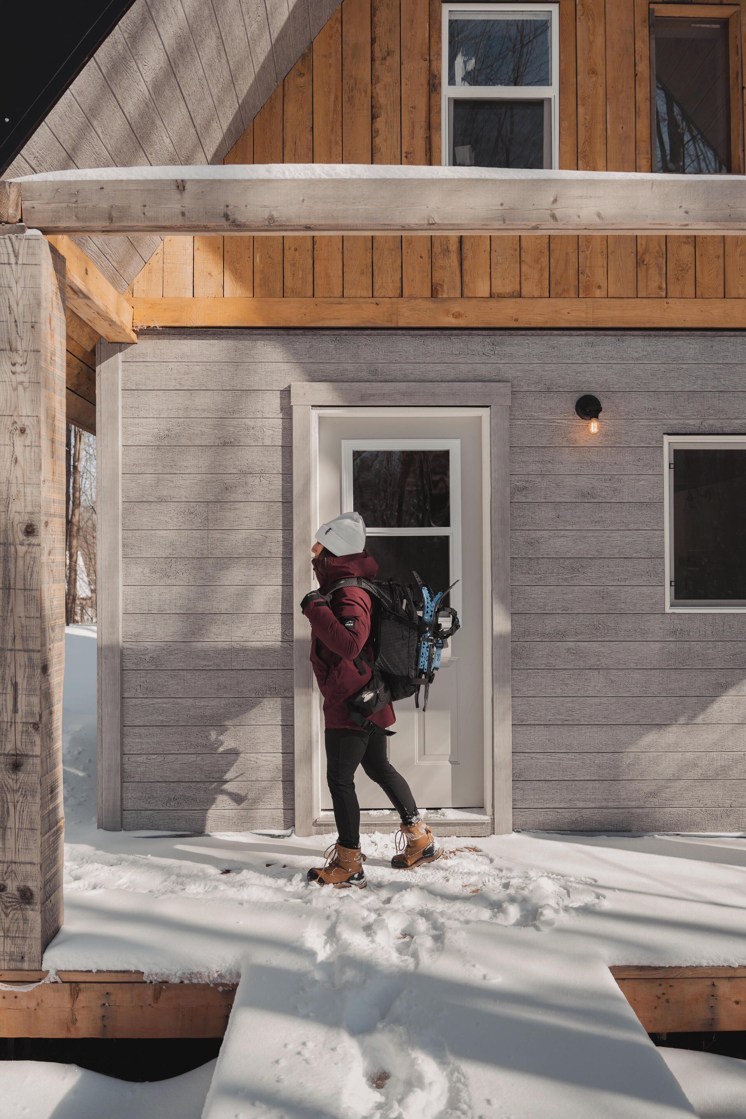A person in winter clothing exiting a wooden house with a snow-covered ground.