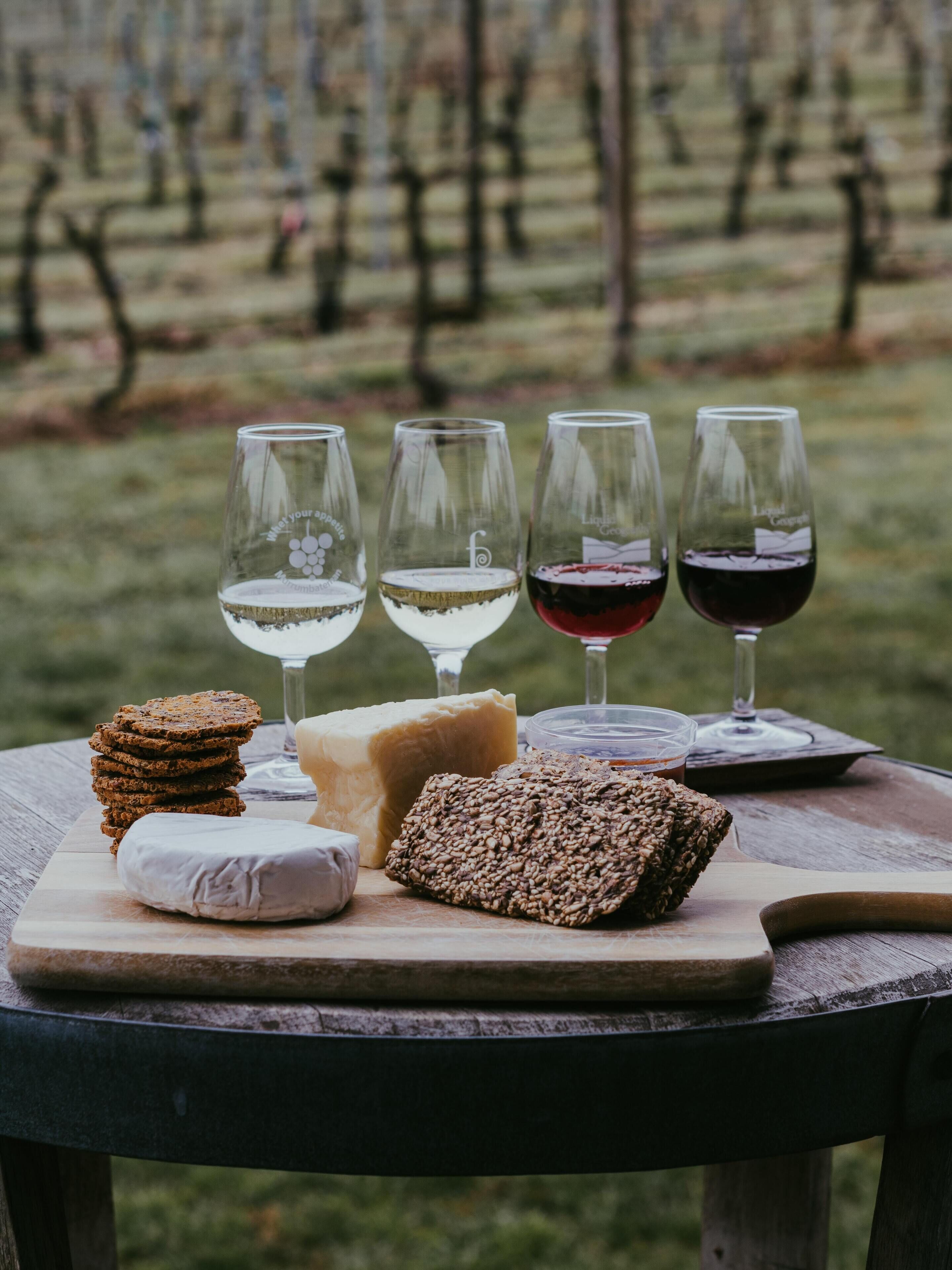 A table with four wine glasses, a stack of crackers, a block of cheese, and a loaf of bread.