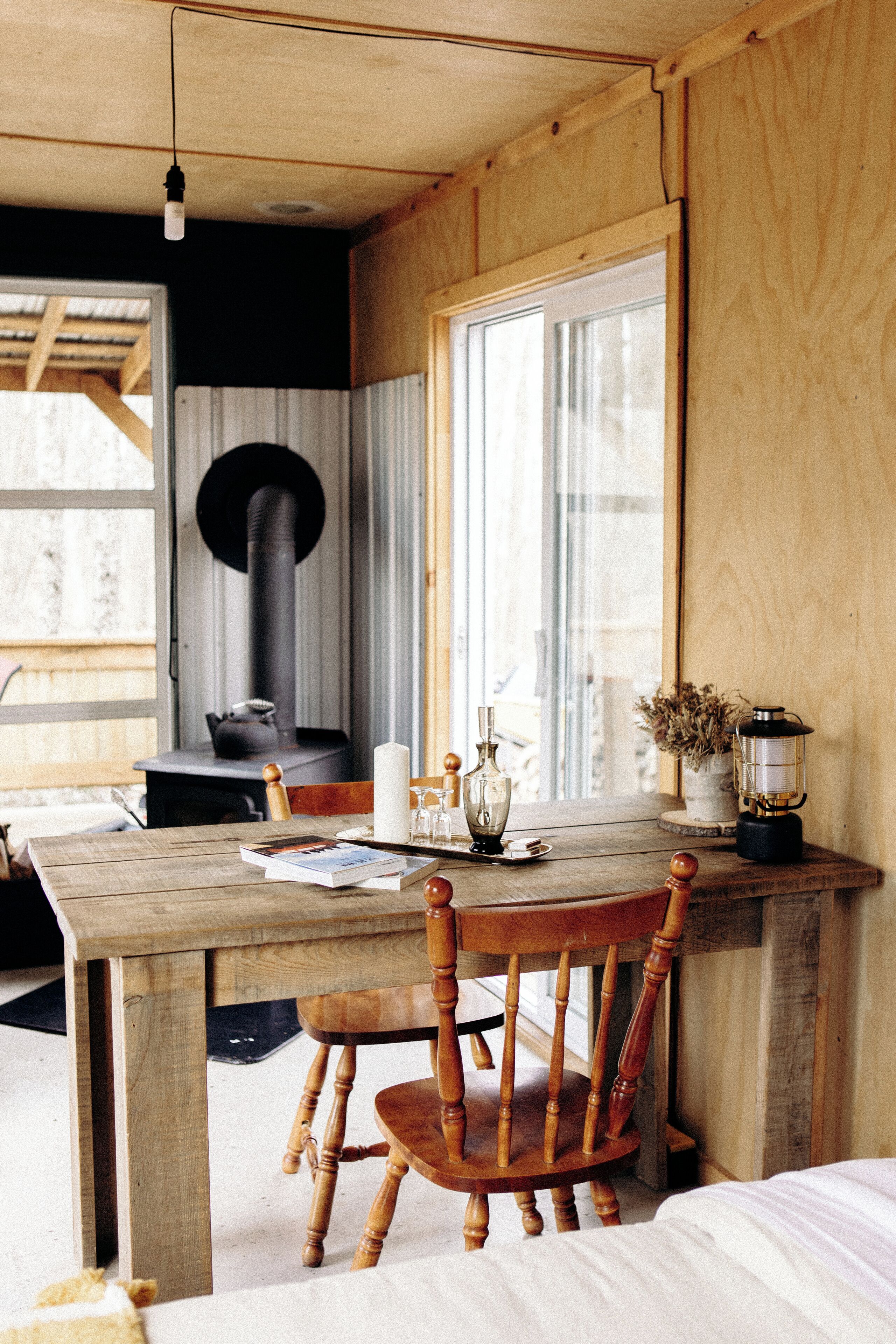 A rustic wooden interior with a large window, a wooden table, a chair, a candle, a glass bottle, and a lantern.