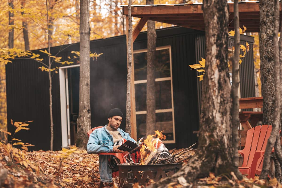 A person standing by a campfire in a wooded area with a small cabin in the background.