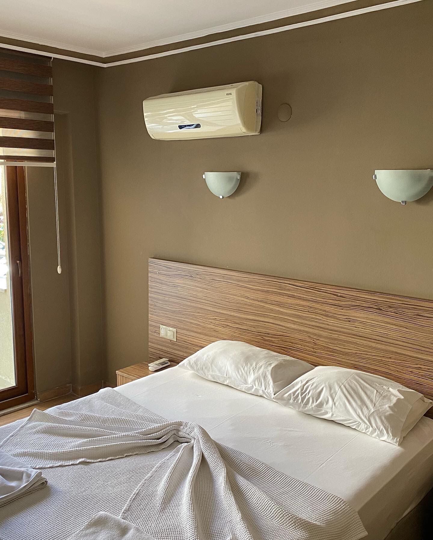 A hotel room with a bed, a wall-mounted air conditioner, two wall sconces, a window with blinds, and a wooden headboard.