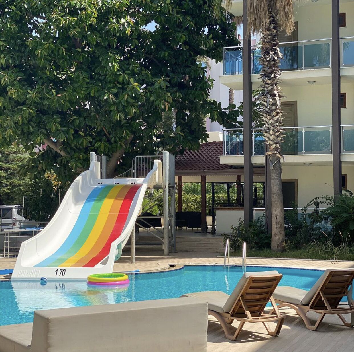 Uma piscina com um escorrega em formato de arco-íris, cadeiras de descanso e uma palmeira.