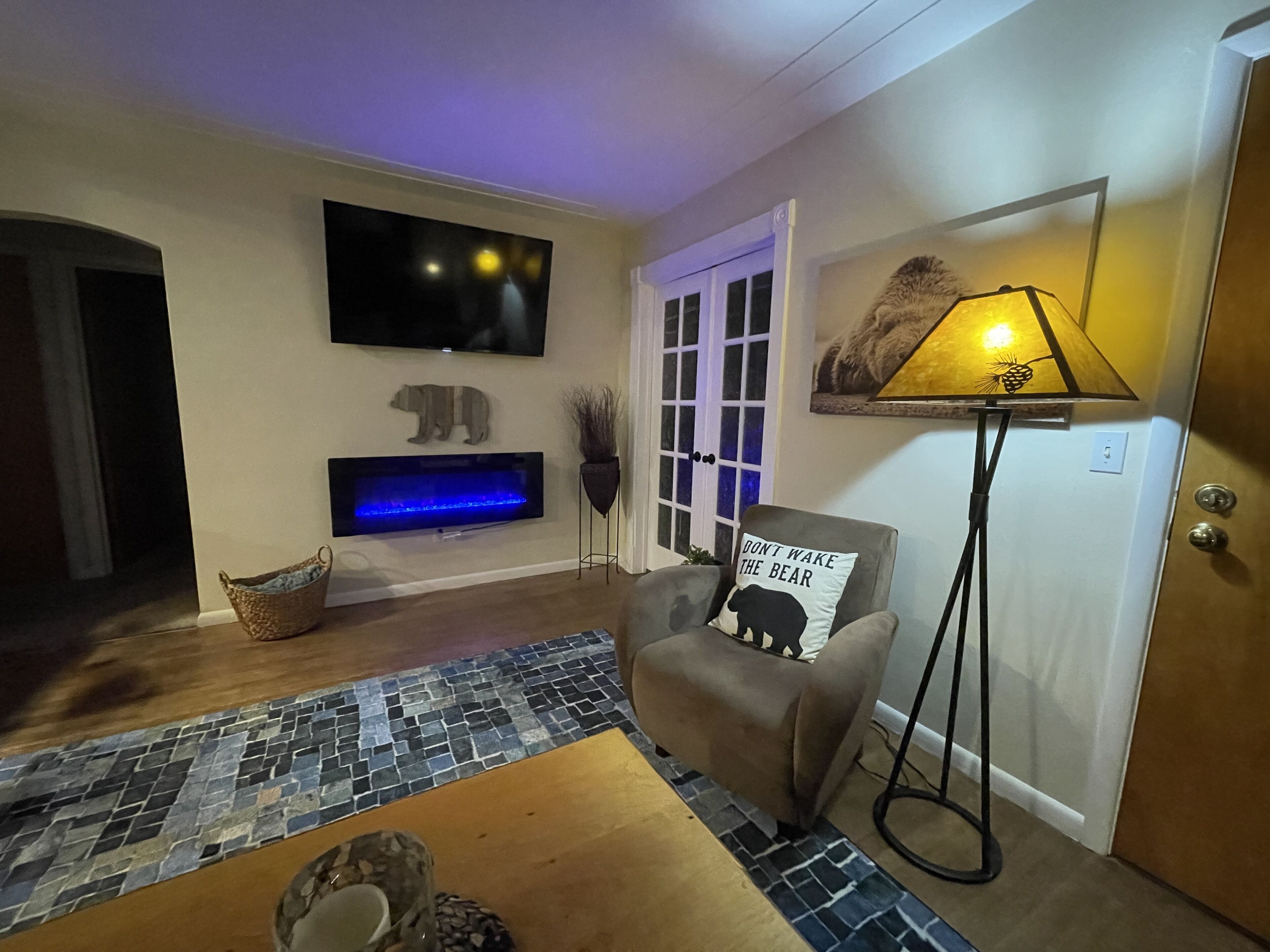 A living room with a flat-screen TV, a fireplace, a chair with a bear pillow, a floor lamp, and a patterned rug.