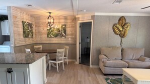 A modern living room with a beige sofa, a dining area with a wooden table and chairs, and a kitchen with white cabinets and a granite countertop.