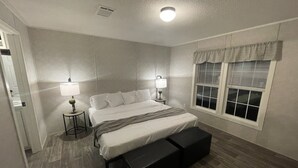 A bedroom with a bed, two bedside tables, a window with blinds, a ceiling light, and a door.
