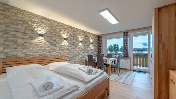 Een slaapkamer met een stenen muur, een houten bed, een eethoek met een tafel en stoelen, en uitzicht door een glazen deur.
