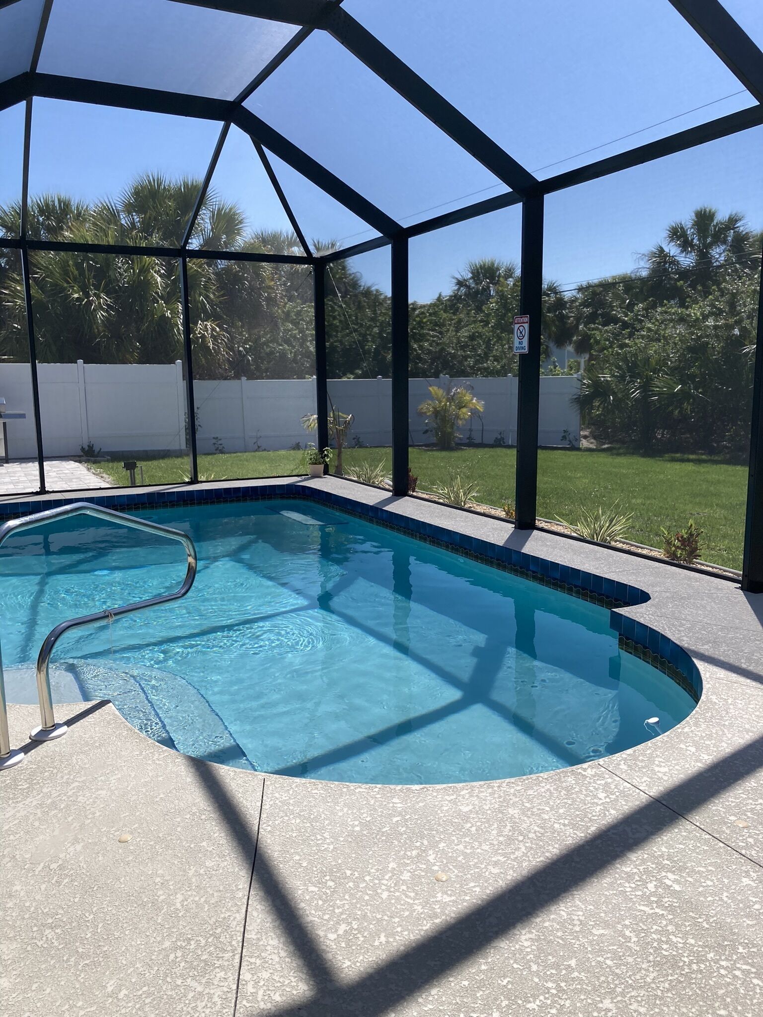 A covered outdoor swimming pool with a clear blue water, surrounded by a white fence and lush greenery.