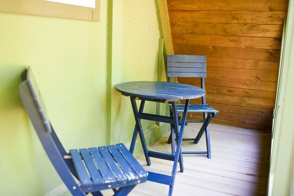 Een klein, afgesloten terras met een houten vloer en een groene wand. Er staan twee blauwe metalen stoelen en een kleine ronde tafel.