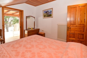 A bedroom with a large bed, wooden dresser, and a mirror. There is a door leading to a balcony with a view of trees and a building.