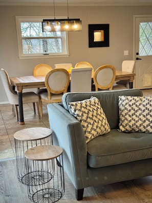 A modern living room with a grey sofa, patterned cushions, a round coffee table, and a dining area with wooden chairs and a table.