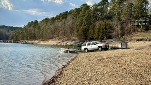 A white SUV parked on a pebbled beach with a dock extending into calm water, surrounded by dense forest and a clear sky.