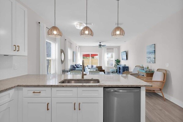 A modern kitchen with white cabinets, a stainless steel sink, and a dishwasher. There are wicker pendant lights, a ceiling fan, and a dining area with a table and chairs.