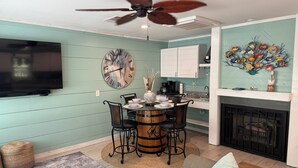 A dining area with a round table, chairs, a ceiling fan, a wall-mounted clock, and a kitchen area with cabinets and a decorative fish mural. - The Pelican Post 1 bedroom beach cottage (Carrabelle)