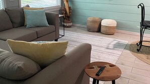 A living room with a sofa, ottomans, a coffee table, a TV, and a window with blinds. - The Pelican Post 1 bedroom beach cottage (Carrabelle)