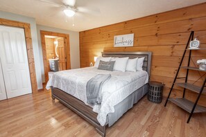 Un dormitorio con cabecera de madera, cama con cubrecama de encaje, mesita de noche con lámpara y un reloj fijado a la pared.