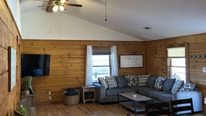 Una habitación acogedora de estilo cabaña, con paredes de madera, ventilador de techo, televisor de pantalla plana montado en la pared, sofá gris con cojines estampados, mesa de comedor de madera con sillas y una mesita auxiliar con una cesta.