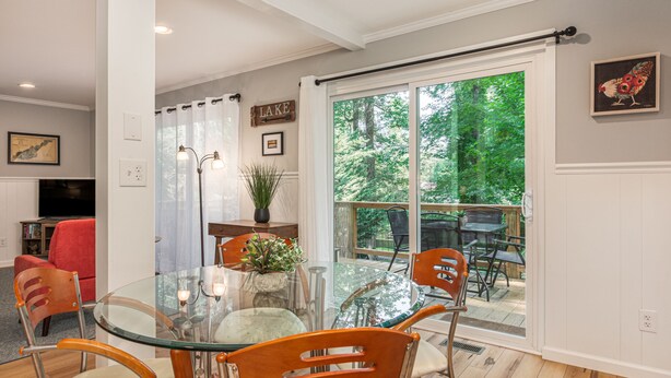 A dining area with a glass table and chairs, a potted plant, and a view of a deck and trees. - Lake Escape - Hot Tub & Lake Access - Pet Friendly (Hendersonville)