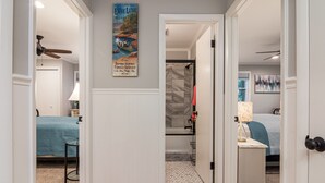 A hallway with a ceiling fan, a poster, a bathroom with a shower, and a bed with a blue quilt. - Lake Escape - Hot Tub & Lake Access - Pet Friendly (Hendersonville)