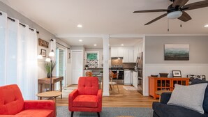 A modern living room with a red armchair, a blue sofa, and a wooden coffee table. There is a ceiling fan and a kitchen in the background. - Lake Escape - Hot Tub & Lake Access - Pet Friendly (Hendersonville)