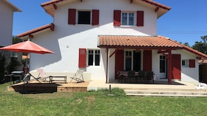 Une maison à deux étages avec un toit recouvert de tuiles, une terrasse couverte avec une table et des chaises, et un parasol rouge.