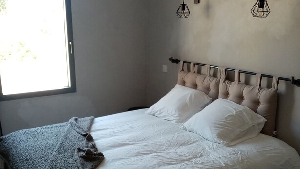 Un lit bien fait, avec du linge de lit blanc et un plaid gris, une fenêtre avec des stores et deux appliques murales.