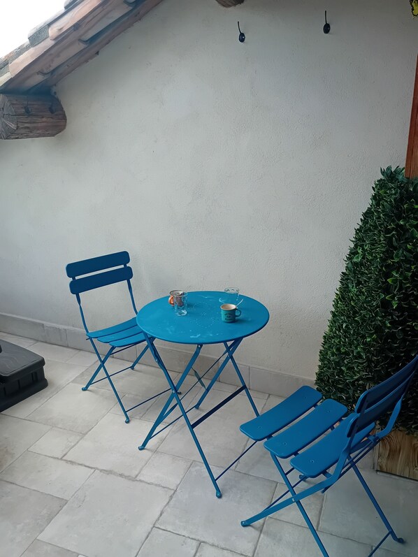 Une table et des chaises en métal bleu sont disposées sur une terrasse carrelée, avec un mur blanc et une petite haie verte en arrière-plan.
