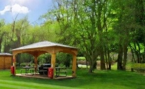 A wooden gazebo with a grill and picnic table in a grassy area surrounded by trees. - Birdsong Lodge Dog Friendly Nr Sutton on Sea (Mablethorpe)