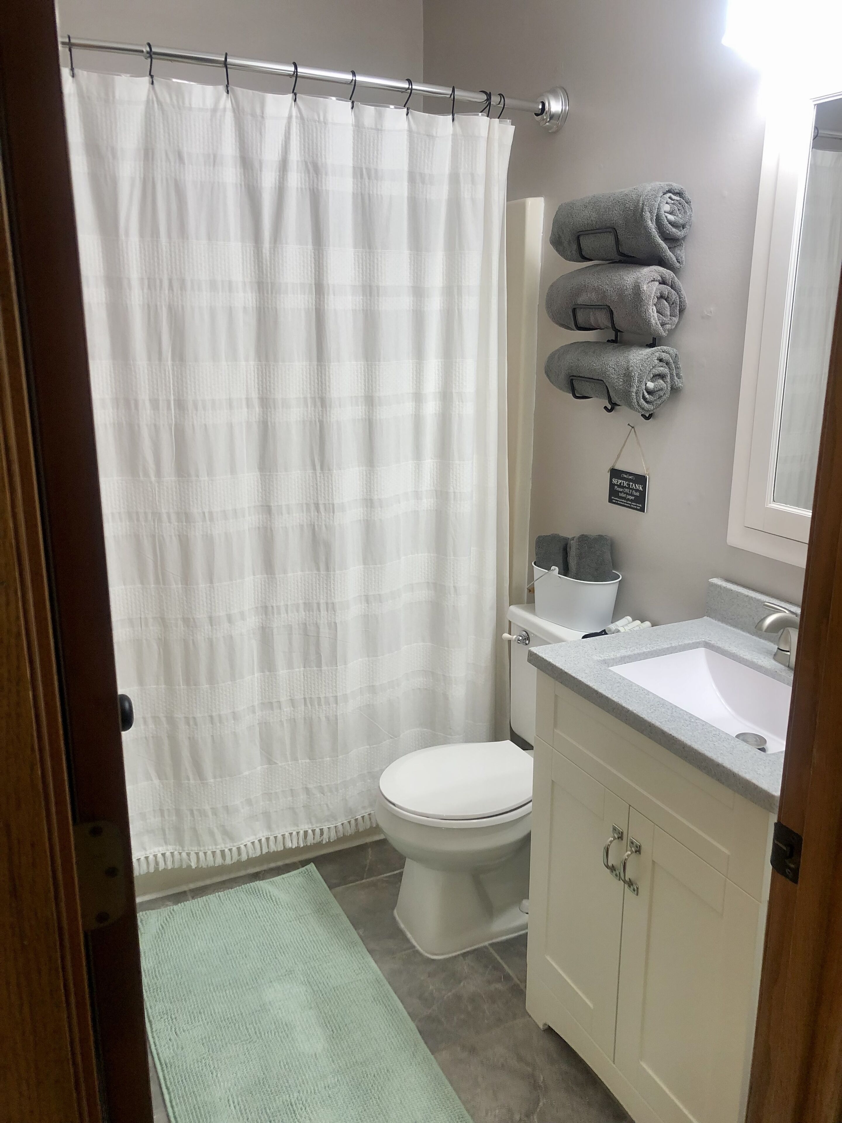 Une salle de bain avec des toilettes blanches, un lavabo et un rideau de douche. Des serviettes sont accrochées au mur, et une pancarte indique : « FOSSE SEPTIQUE ».