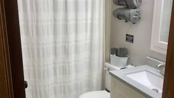 Une salle de bain avec des toilettes blanches, un lavabo et un rideau de douche. Des serviettes sont accrochées au mur, et une pancarte indique : « FOSSE SEPTIQUE ».