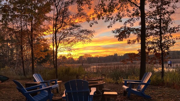 Un terrain de camping équipé de chaises Adirondack, d’un foyer et offrant une vue sur le coucher de soleil au-dessus d’un champ.