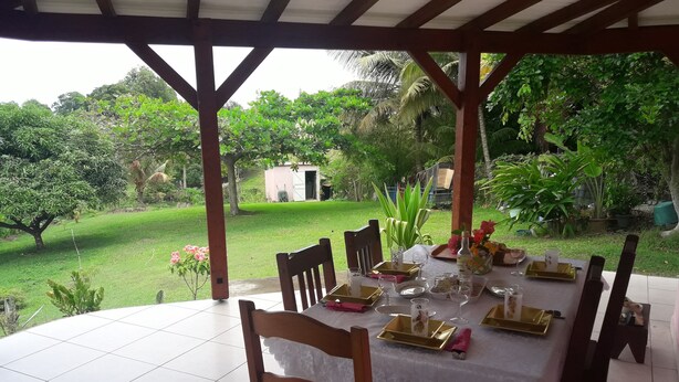 A covered outdoor dining area with a table set for four, surrounded by chairs, and a view of a lush garden. - Villa of 100 m2 flower gardens and fruit trees (Le Moule)