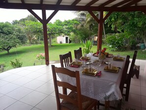 A covered outdoor dining area with a table set for four, surrounded by chairs, and a view of a lush garden. - Villa of 100 m2 flower gardens and fruit trees (Le Moule)