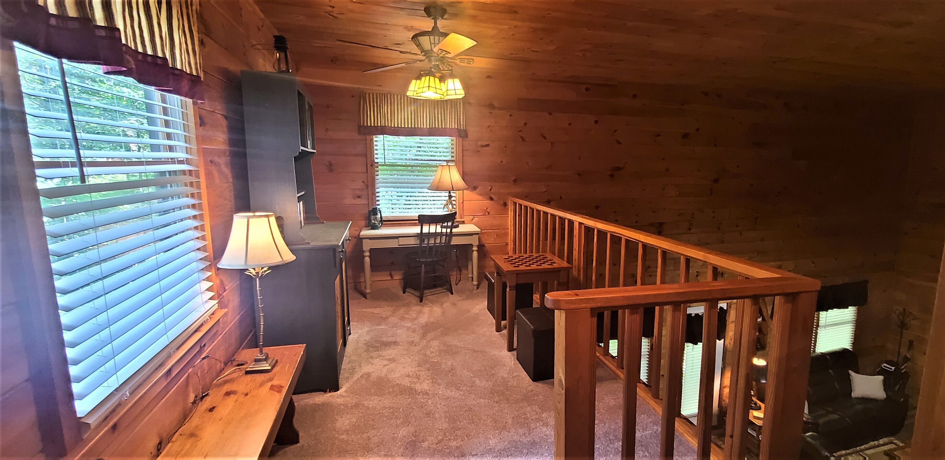 A cozy cabin interior with a wooden staircase, a desk with a lamp, and a view through the window.