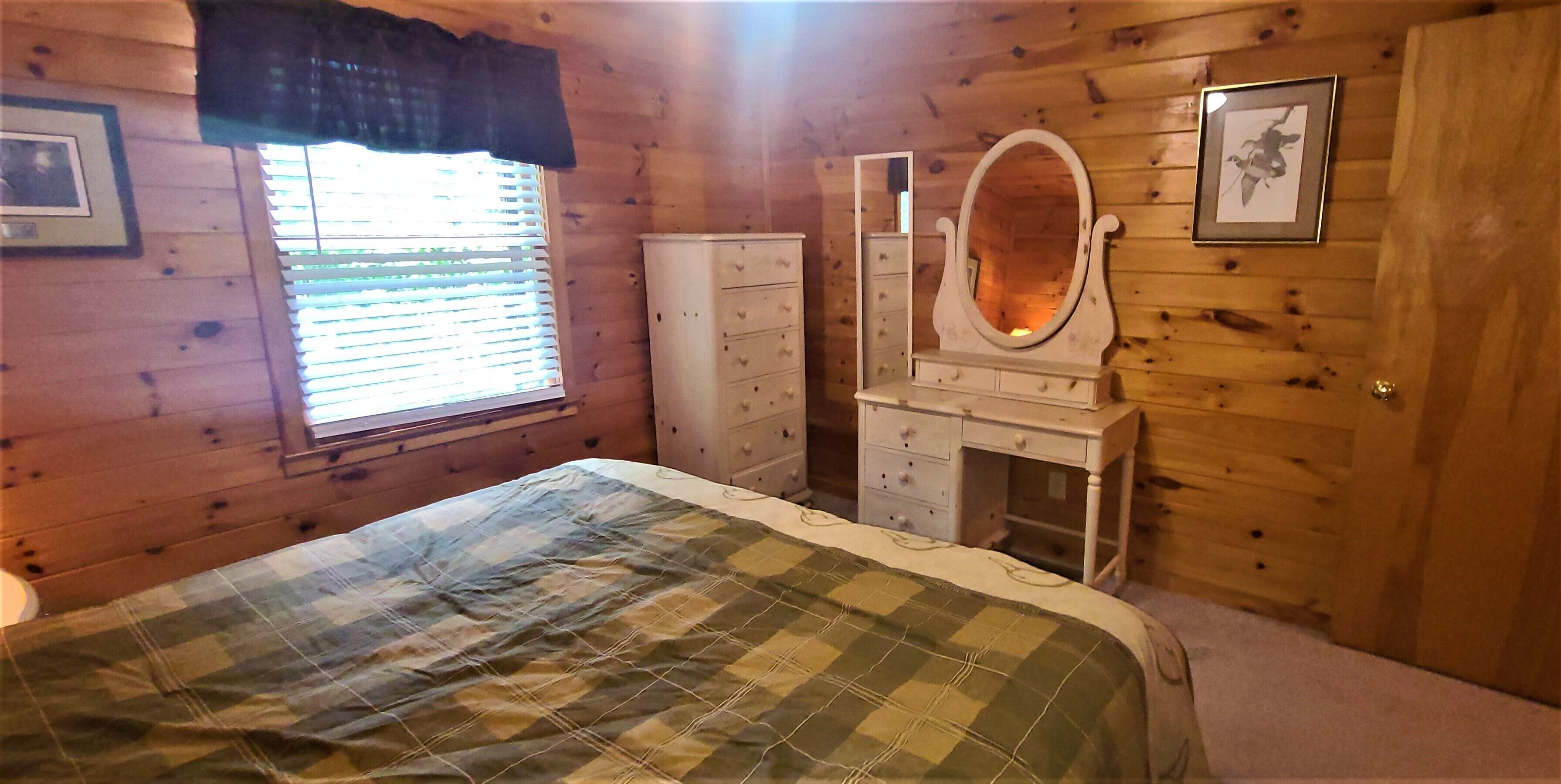 A wooden cabin bedroom with a bed, dresser, vanity with mirror, and window with blinds.